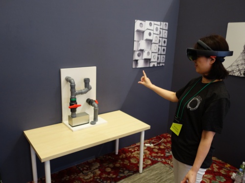 「Microsoft Remote Assist」のデモを体験している様子。HoloLensを着けた体験者が、空間に表示された写真やビデオ通話のウインドウをハンドジェスチャーで操作し、パイプについた2つのオレンジ色の栓を操作しようとしている