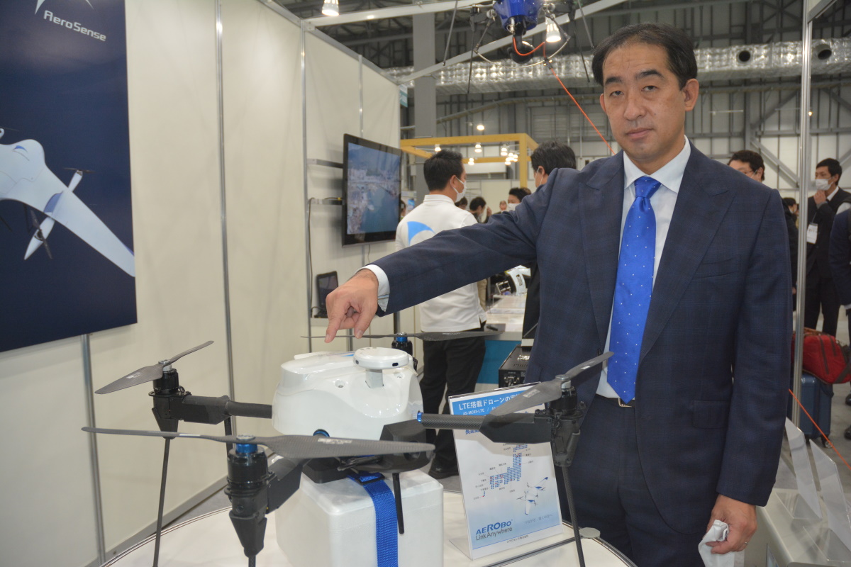回転翼ドローン「エアロボAS-MC03-LTE」。2021年11月24～26日の「第7回国際ドローン展」に初めて出展した。写真右はエアロセンスの佐部浩太郎社長。指でさす箇所に、SIMカードを挿入したLTE通信モジュールを内蔵している（写真：日経クロステック）