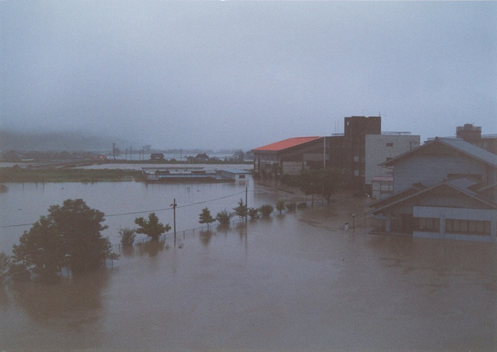 1990年夏の大水害で被災した、建て替え前の佐賀県嬉野市立塩田中学校付近の様子。右奥に見える赤い屋根や茶色の建物が体育館や校舎だ。大水害の際に同校周辺は1m程度浸水した。旧校舎はこの際、床上浸水の被害を受けた（写真：嬉野市教育委員会）