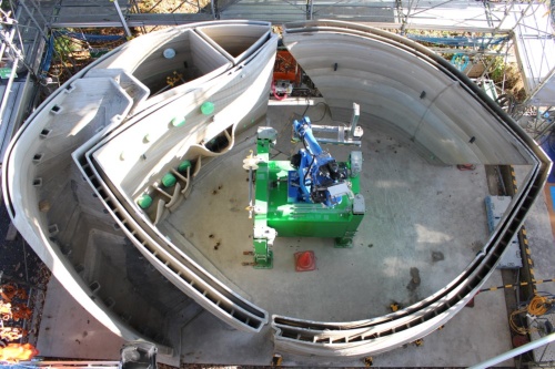 建設中の施設を真上から見た様子。施設の内部にロボットアーム型の建設3Dプリンターを据え付けて印刷していく。印刷場所に応じて、緑色の台座ごとプリンターを盛り替える。写真左側の屈曲部が階段で、その奥が機械室だ(写真:大林組)