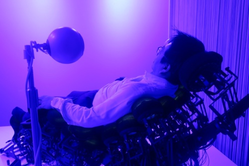 Media Ambition Tokyo 2019で、共感覚体験装置「Synesthesia X1-2.44」を体験する筆者｡黒い椅子は44個の振動子をつなげて作っている。椅子の脇にある丸い装置はスピーカー
