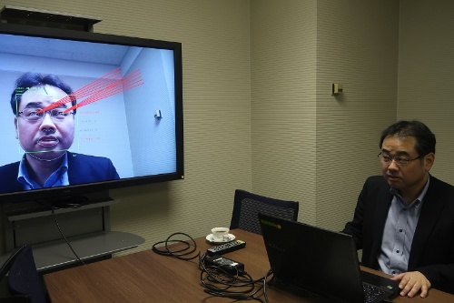 NECが開発した「遠隔視線推定技術」。視線の方向を目から出る赤い直線で見える化している。緑色の枠は、人の顔と目の検出部分。今回は手前のノートパソコンに取り付けたカメラで、パソコンの前に座る人の視線を検知している