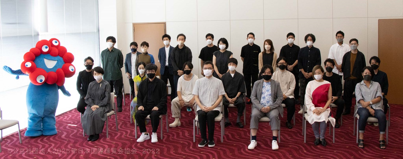 選ばれた若手建築家20組、合計23人と評価委員（中央前列の3人）、そして万博の公式キャラクター「ミャクミャク」（左）（写真：2025年日本国際博覧会協会）