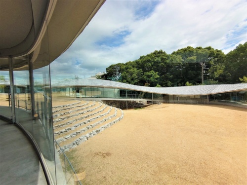 石段を下りた右側は多目的広場。施設のうねる屋根がよく見える。屋根材には、地元の庵治(あじ)石の瓦を約3万4000枚使った。庵治石は高級な墓石として有名だが、表面がきれいな部分しか使われないため端材が大量に出る。それを屋根材に利用することを周防氏が思いついた(写真:日経クロステック)