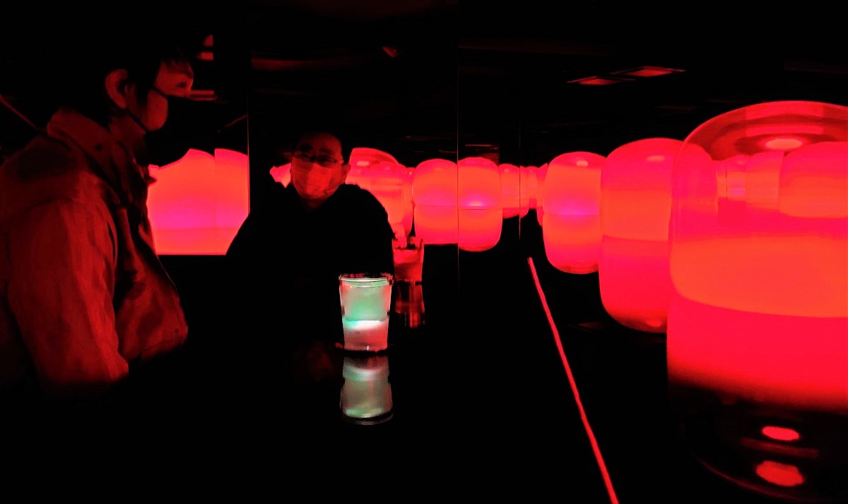 2週間ぶりに外出した猪子氏と、新作「TeamLab：Tea Time in the Soy Sauce Storehouse」の前で話をする私（右）。地下は真っ暗で、赤く光る無数のランプが水盤に浮かぶ光景とお茶のグラスだけが見える（写真：日経クロステック）