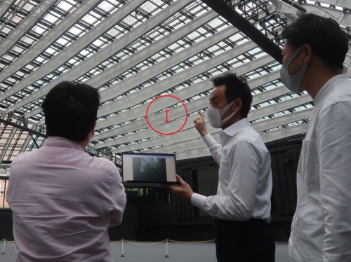 日建設計の設計担当者2人と白い梁を見上げながら、説明を受ける私(左)。中央がファサードエンジニアの村上博昭氏。梁のデプス(上弦材と下弦材の間隔)は最大で1.8mある。写真の赤色矢印の部分(写真:日経クロステック)