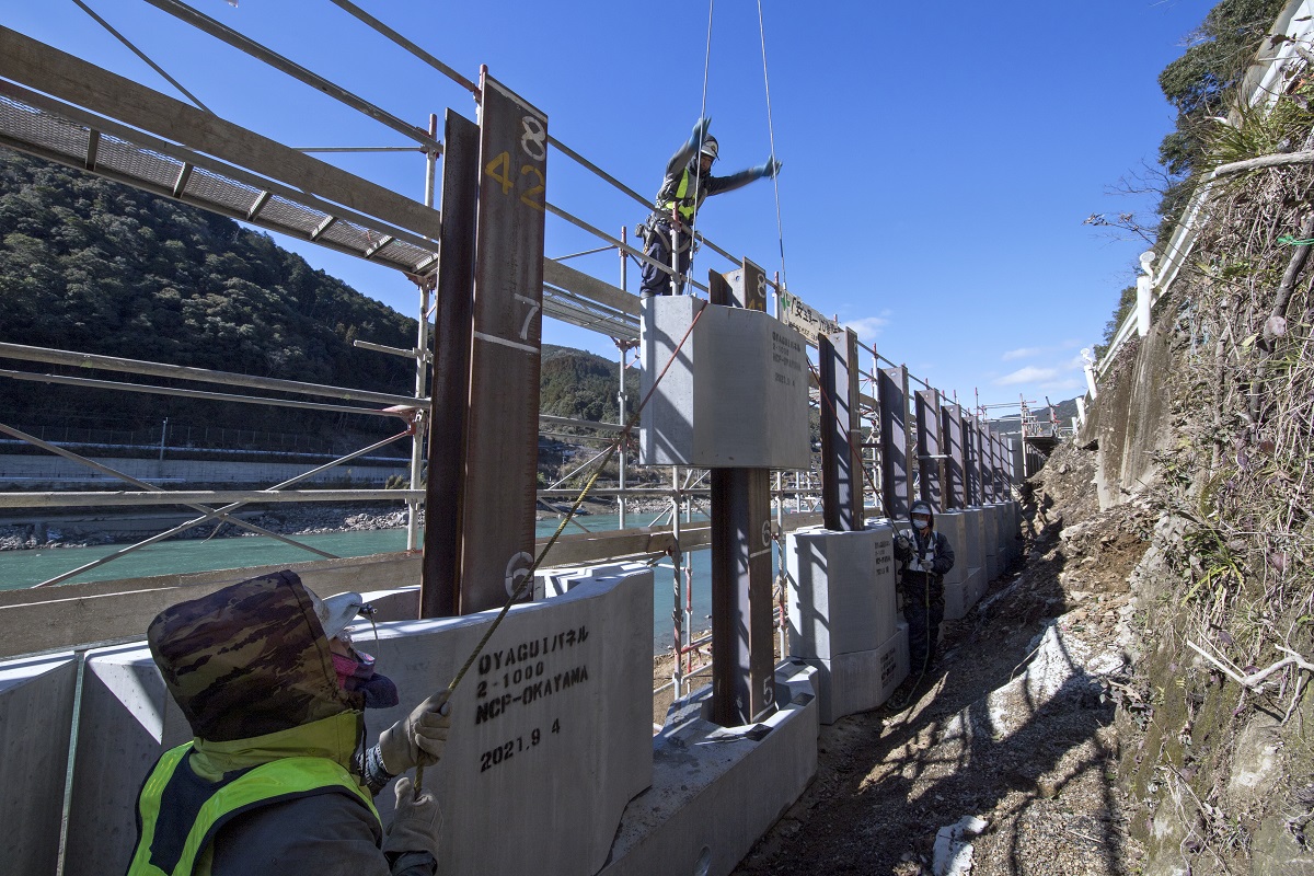 日特建設が味岡建設の1次下請けとして「親杭パネル工法」を施工する熊本県球磨村神瀬地区の現場。写真は鉄筋コンクリート製のプレキャストパネルの位置を調節する様子（写真：イクマ サトシ）