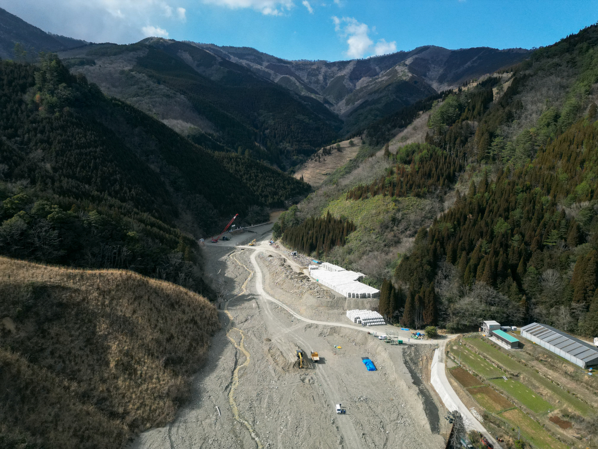 現場は急峻な山間部に位置する。写真中央付近に遠隔地から操作するバックホーなどが見える。治山ダムは写真奥側に設置する（写真：旭建設）