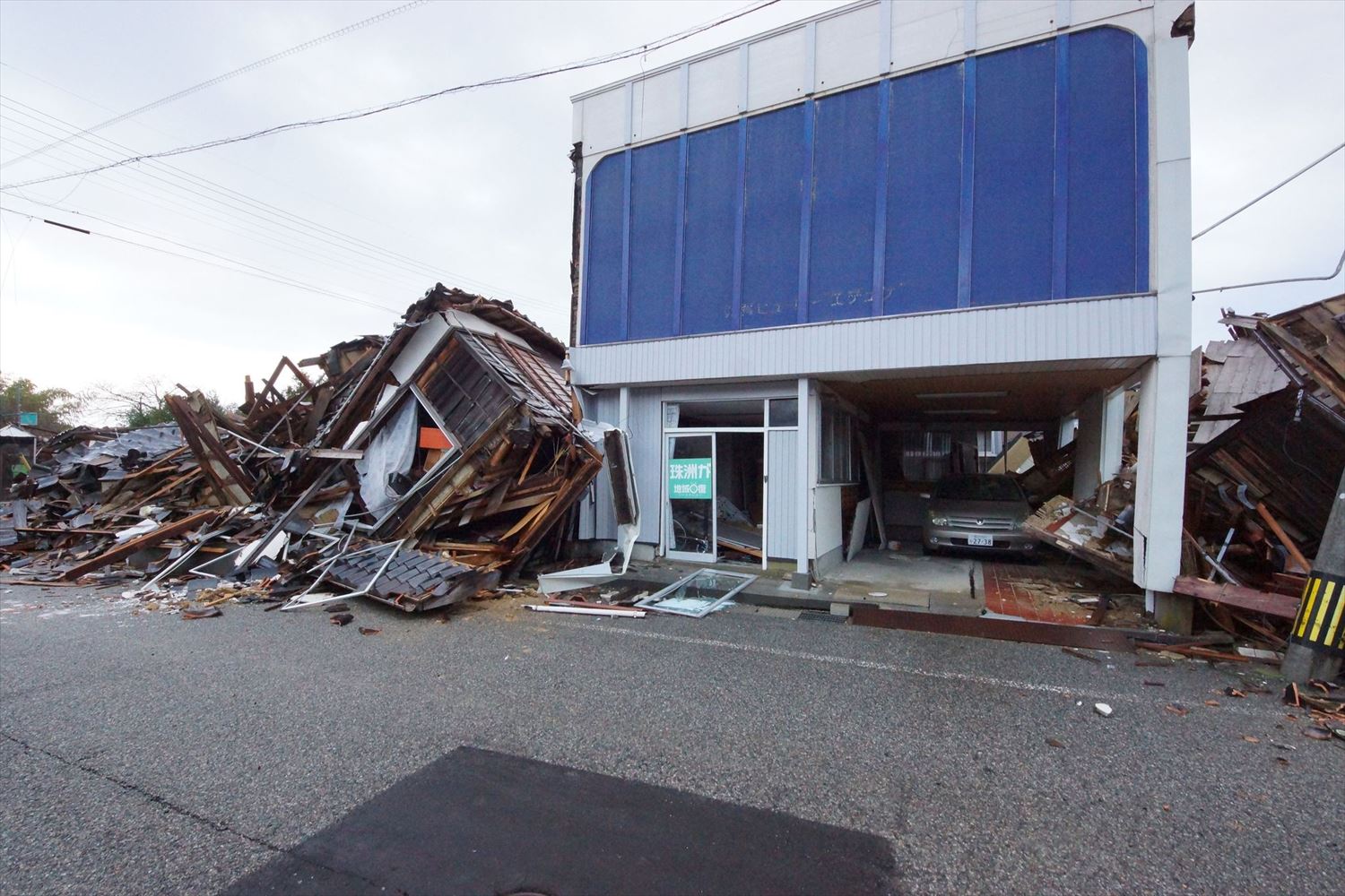 同事務所の24年能登半島地震後の状態。奥能登地震後に入り口周りの耐力壁と、内部の方杖を追加する耐震改修を実施していた。外装材の留め具も締め直していたため、多少の変形に留まった。奥能登地震の時は残っていた左右の木造家屋は、1階部分がすべて潰れる層崩壊が起こった（写真：建物修復支援ネットワーク）