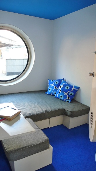 入り口からベッド方向を見る。白い壁に、天井と床の青が映える。オレンジや黒などのカプセルも用意されていた。手前に置かれたスツールを右側の壁ぎわに移動させると、ベッドと一体化したソファになる（写真：守山 久子）