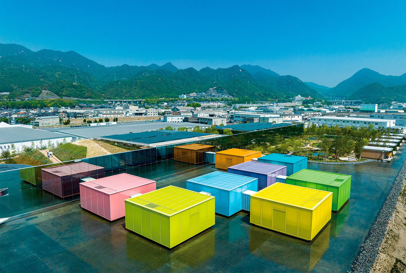 色とりどりの「可動展示室」越しに、企画展示室や管理棟の方向を見る。西側に立つショッピングセンターの目隠しとして、ミラーガラスのスクリーンを南北方向に立てた。写真左側の丘に登ると、可動展示室を上から見下ろすこともできる。施設名の「SIMOSE」は地名ではなく、発注者である丸井産業の下瀬ゆみ子代表の名字からとった（写真：平井 広行）
