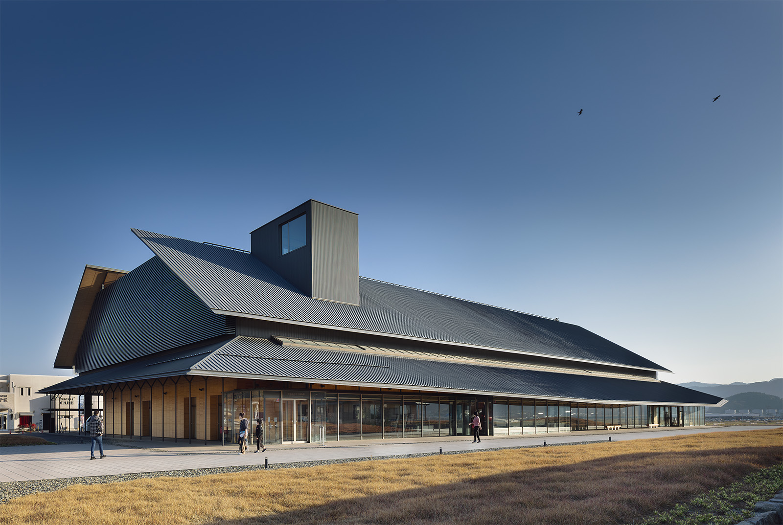 2022年11月5日に開館した陸前高田市立博物館を北西から見る。写真左側のガラス部分が出入り口。ハの字に架かる2枚の大屋根と、1階の上部を取り巻く「裳階（もこし）」のような庇が特徴的。屋根から飛び出しているのは屋上に出るエレベーターの塔屋。屋根や上部の外壁はチタン亜鉛合金板、1階外壁は岩手県産のスギ小幅板パネル（写真：吉田 誠）