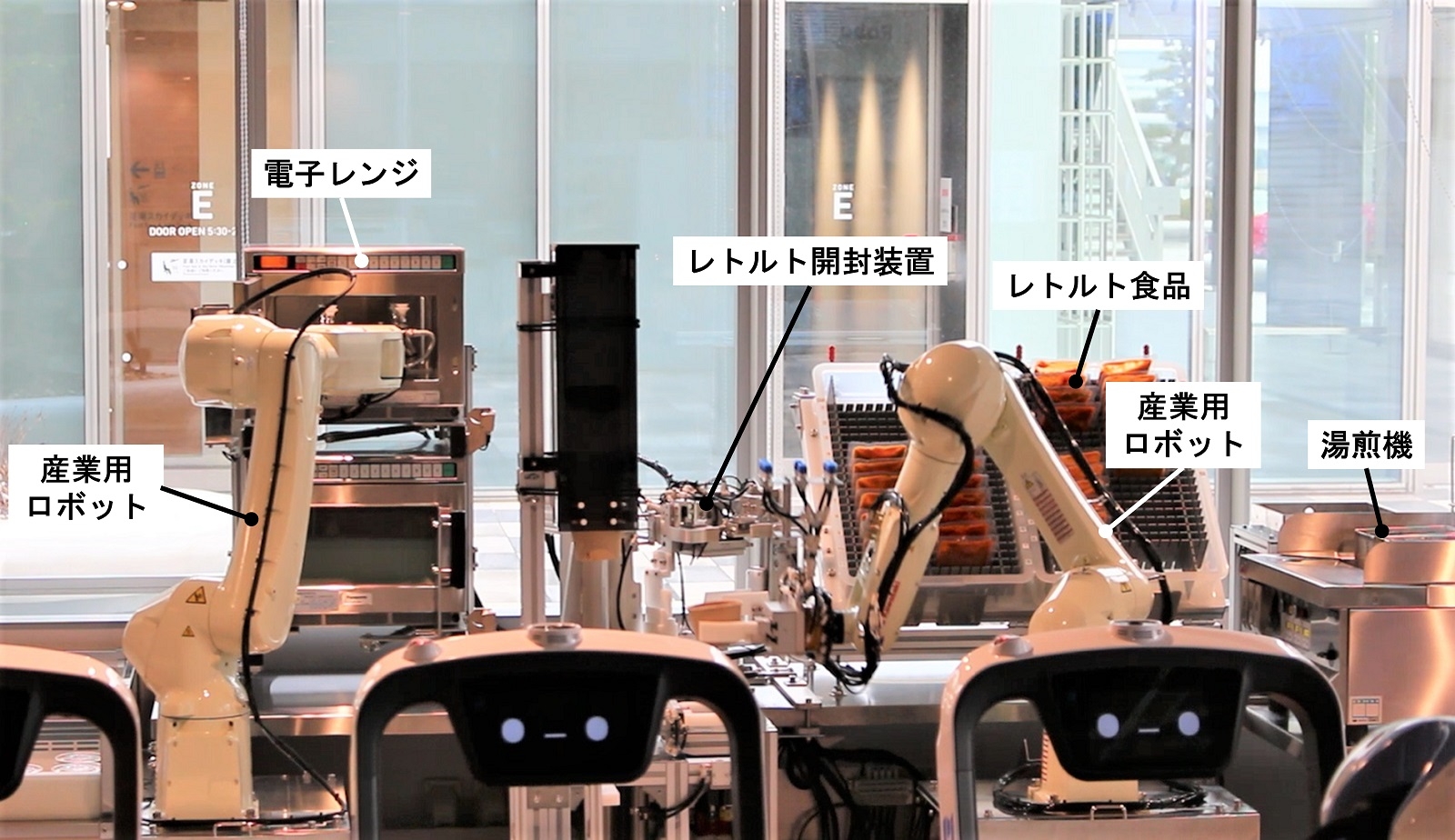 図2　ロボットが料理を準備する様子 左のロボットは、あらかじめ調理された料理を電子レンジで加熱してトレーに載せる。右のロボットは、レトルト食品を湯煎し、レトルト食品を開封する別の装置にセットする。その後、容器に移されたレトルト食品を右のロボットがトレーに載せる。（出所：日経クロステック）