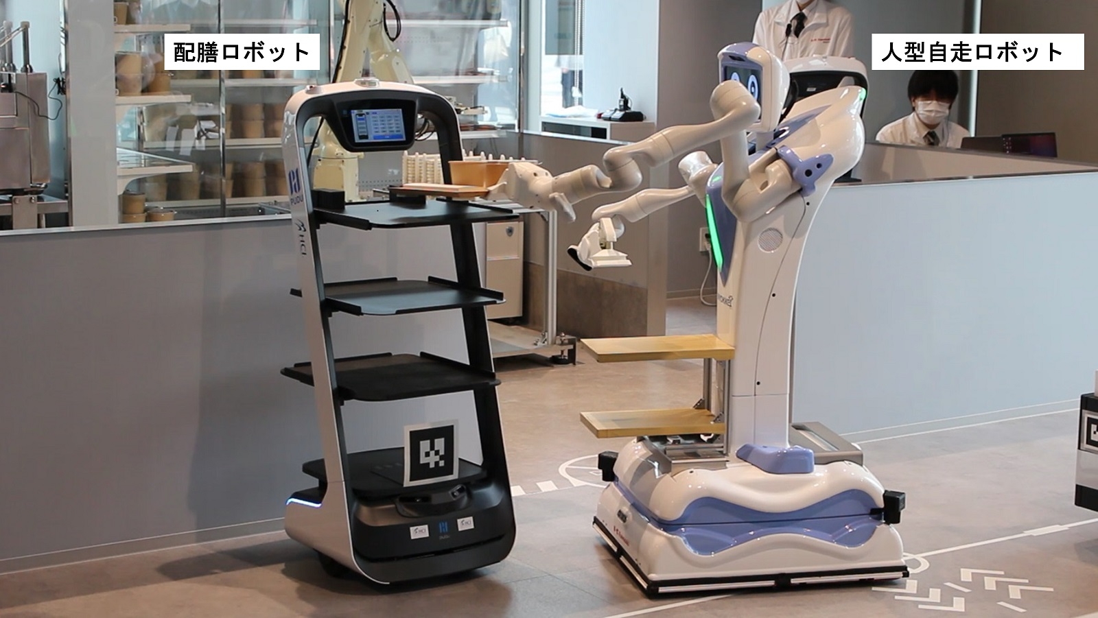 図4　配膳ロボットから人型自走ロボット「Nyokkey」が料理を受け取る様子 配膳ロボットは、写真奥の厨房から飲食エリアまで運ぶ。そこから先は、人型自走ロボットが利用客まで料理を届ける。（出所：日経クロステック）
